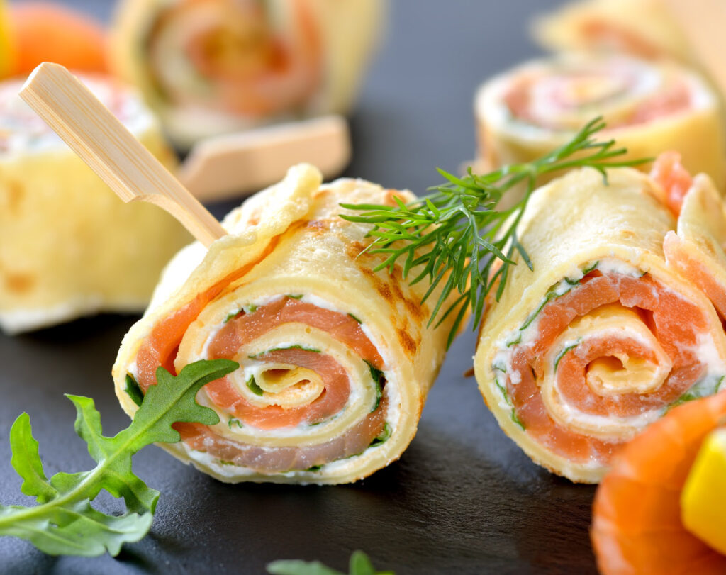 Nahaufnahme von herzhaft gefüllten Crêpe-Röllchen mit Lachs, Frischkäse und Kräutern, garniert mit Dill und Rucola.