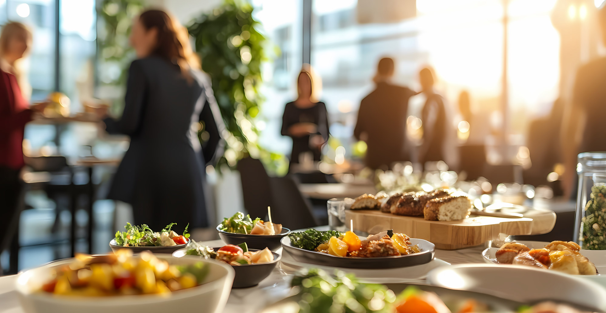 Angerichtete Speisen stehen auf einem Tisch in einem hellen Raum, während im Hintergrund mehrere Menschen bei einer Veranstaltung zusammenstehen.