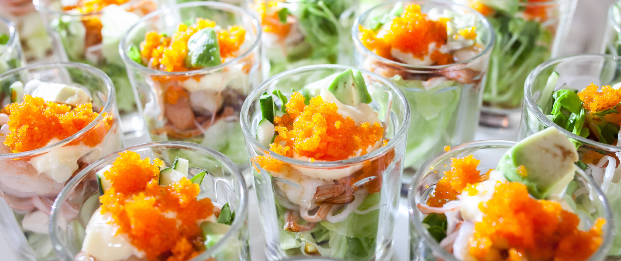 Mehrere kleine Gläser mit angerichteten Vorspeisen aus Salat, Meeresfrüchten, Creme und orangefarbener Garnitur stehen dicht beieinander.