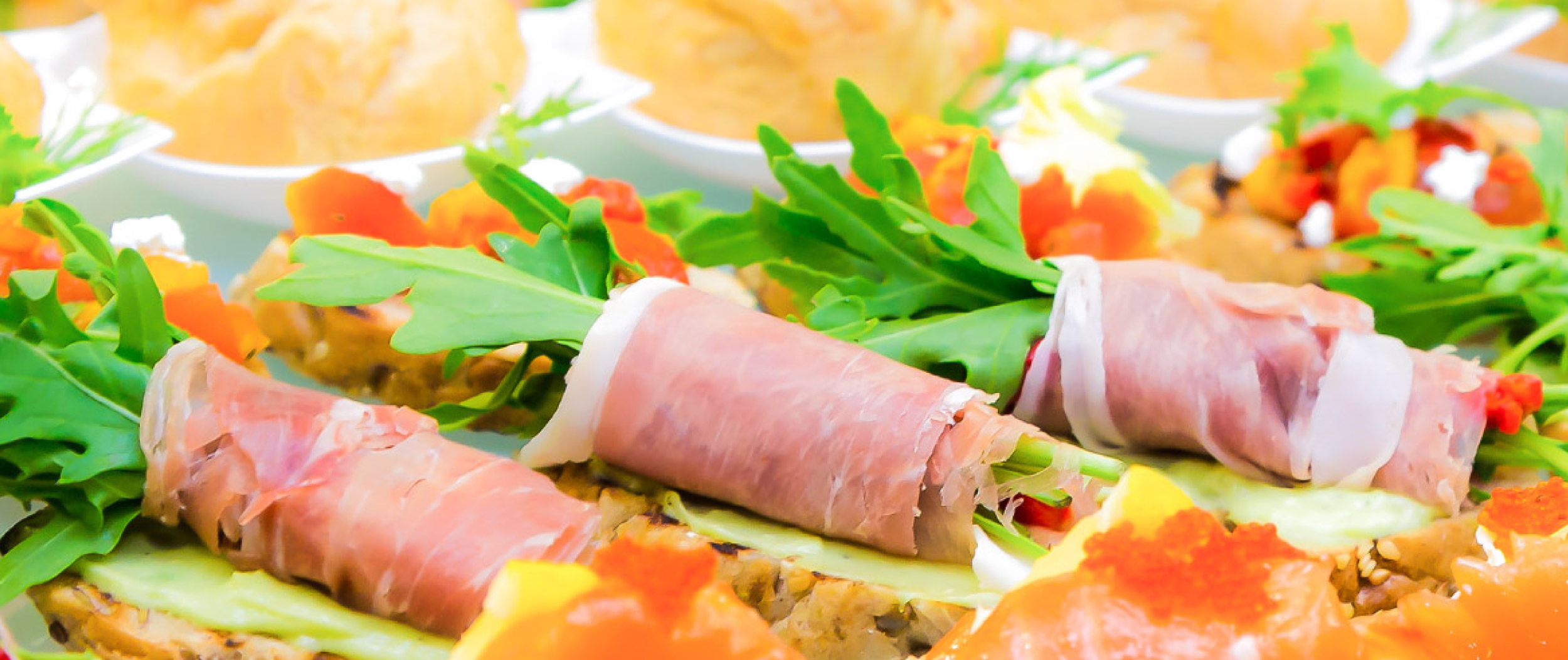 Nahaufnahme von belegten Häppchen mit Schinken, Rucola, Gurke und bunten Garnituren auf einer Platte.