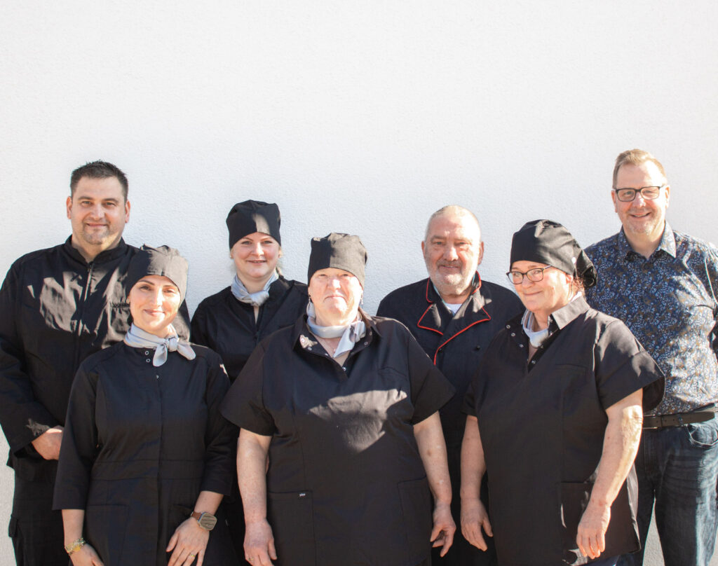 Gruppenfoto eines Catering-Teams vor einer hellen Wand; mehrere Mitarbeitende in schwarzer Arbeitskleidung stehen neben zwei Personen in Alltagskleidung.